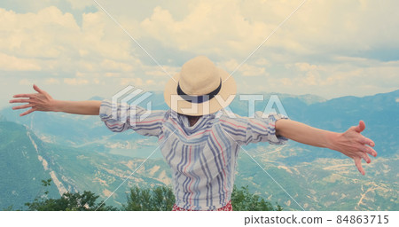 Happy traveler arms wide open to sides on mountain top. Unrecognizable women in hat hugs mountains landscape with fluffy clouds. Travel, trekking concept. Happy traveler enjoys mountain valley view 84863715