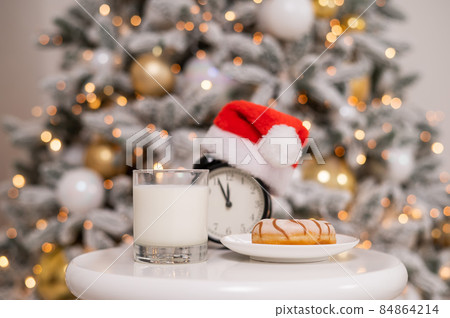 Alarm clock in a santa claus hat with a glass of milk and a donut on the background of a christmas tree. Alarm clock in a santa claus hat with a glass of milk and a donut on the background of a christmas tree. 84864214