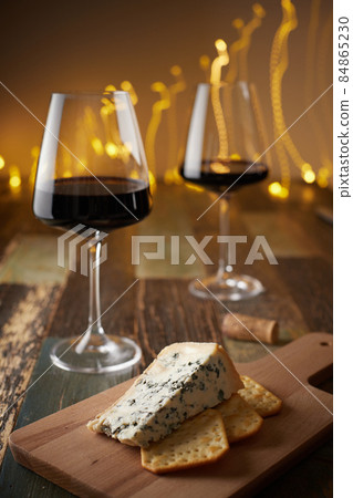 Still life of two glasses of wine, cheese with blue mold and crackers on a wooden board against the background of a New Year's garland. Front view. 84865230