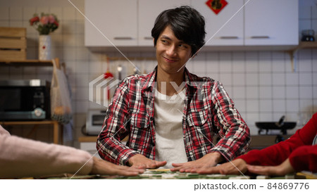 son looking at his family with sly smile while shuffling tiles on mahjong table. indoor recreation for chinese lunar new year's eve concept. translation: luck 84869776