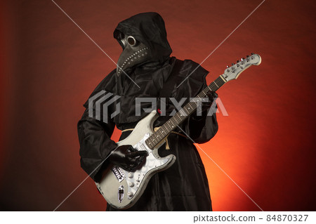 A man in a plague doctor costume plays the guitar emotionally on a red background 84870327