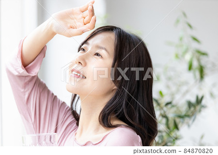 A woman in room wear standing by the window with a glass of water 84870820