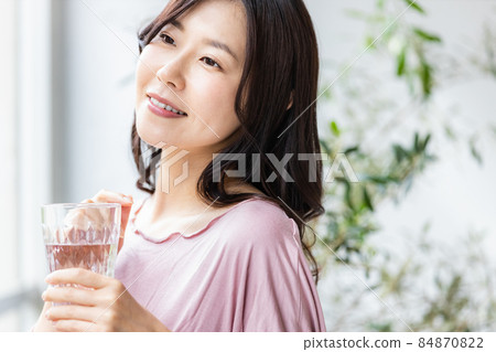 A woman in room wear standing by the window with a glass of water 84870822