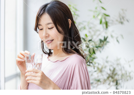 一個身著房間的女人站在窗邊，拿著一杯水 84870823