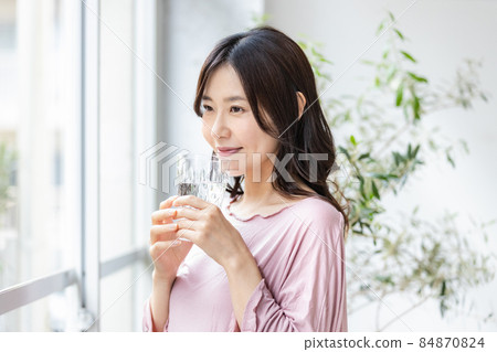 A woman in room wear standing by the window with a glass of water 84870824