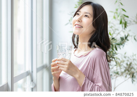 A woman in room wear standing by the window with a glass of water 84870825