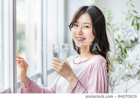 A woman in room wear standing by the window with a glass of water 84870829