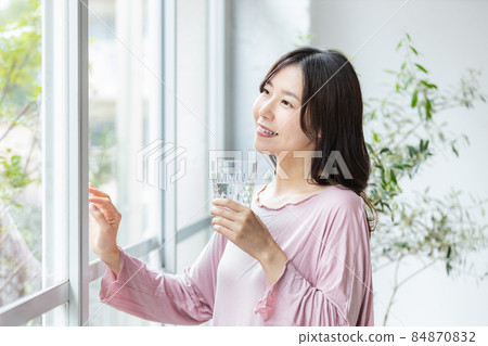A woman in room wear standing by the window with a glass of water 84870832