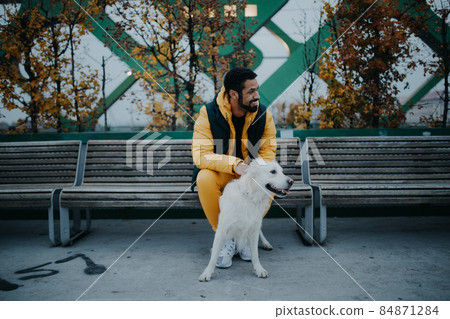 Happy young man sitting on bench and holding his dog outdoors in town in autumn. 84871284