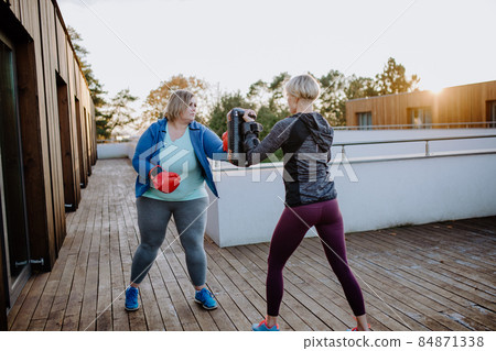 Overweight woman training boxing with personal trainer outdoors on terrace. 84871338
