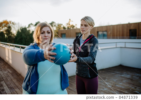 Overweight woman holding ball and exercising with personal trainer in outdoors on gym terrace. 84871339