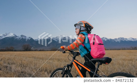 One caucasian children walk with bike in wheat field. Little girl walking black orange cycle on background of beautiful snowy mountains. Biker stand with backpack and helmet. One caucasian children walk with bike in wheat field. Little girl walking black orange cycle on background of beautiful snowy mountains. Biker stand with backpack and helmet. 84872132