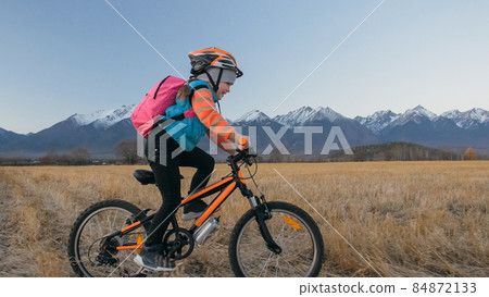One caucasian children rides bike in wheat field. Little girl riding black orange cycle on background of beautiful snowy mountains. Biker motion ride with backpack, helmet. Mountain bike hardtail. One caucasian children rides bike in wheat field. Little girl riding black orange cycle on background of beautiful snowy mountains. Biker motion ride with backpack, helmet. Mountain bike hardtail. 84872133