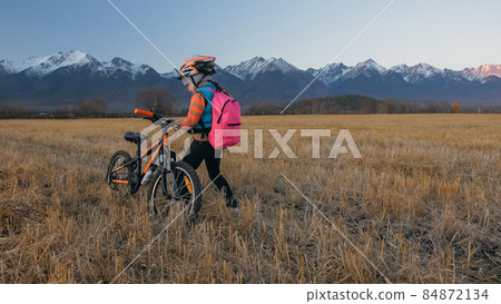 One caucasian children walk with bike in wheat field. Little girl walking black orange cycle on background of beautiful snowy mountains. Biker stand with backpack and helmet. One caucasian children walk with bike in wheat field. Little girl walking black orange cycle on background of beautiful snowy mountains. Biker stand with backpack and helmet. 84872134