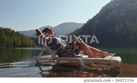 Woman lie on a sunbed in sunglasses and a boho silk shawl. Girl rest on a flood wood underwater pier. The pavement is covered with water in the lake. Woman lie on a sunbed in sunglasses and a boho silk shawl. Girl rest on a flood wood underwater pier. The pavement is covered with water in the lake. 84872154