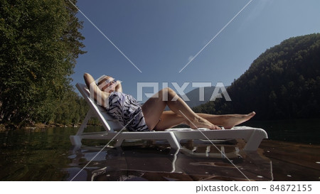 Woman lie on a sunbed in sunglasses and a boho silk shawl. Girl rest on a flood wood underwater pier. The pavement is covered with water in the lake. 84872155