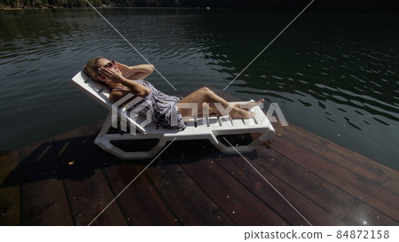Woman lie on a sunbed in sunglasses and a boho silk shawl. Girl rest on a flood wood underwater pier. The pavement is covered with water in the lake. Woman lie on a sunbed in sunglasses and a boho silk shawl. Girl rest on a flood wood underwater pier. The pavement is covered with water in the lake. 84872158