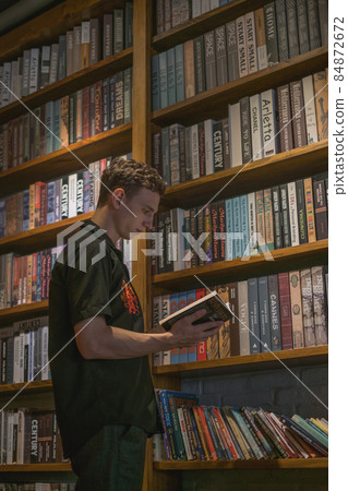 Young caucasian man standing near the wooden shelves with books in the cafe. Holding and reading the book. Vintage style in a cafe. Library. Young caucasian man standing near the wooden shelves with books in the cafe. Holding and reading the book. Vintage style in a cafe. Library. 84872672