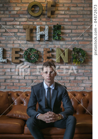 Portrait of young caucasian business man in formal suit sitting on brown leather sofa. Handsome manager sitting on a couch in an open space in office Portrait of young caucasian business man in formal suit sitting on brown leather sofa. Handsome manager sitting on a couch in an open space in office 84872673