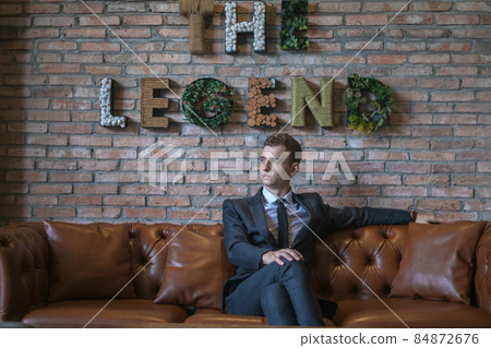 Portrait of young caucasian business man in formal suit sitting on brown leather sofa. Handsome manager sitting on a couch in an open space in office Portrait of young caucasian business man in formal suit sitting on brown leather sofa. Handsome manager sitting on a couch in an open space in office 84872676