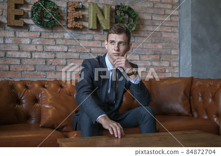 Portrait of young caucasian business man in formal suit sitting on brown leather sofa. Handsome manager sitting on a couch in an open space in office 84872704