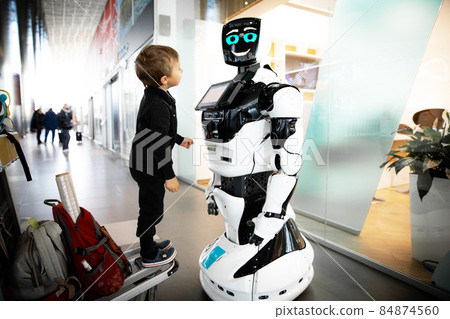 Small boy of European appearance in a black overalls stands on a trolley for suitcases and looks with interest at a robot that is standing and smiling in a public place. 84874560