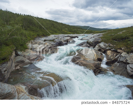 Milky blue glacial Laddejahka river rapids with colorful stones in Lapland landscape with green mountains and birch trees at Padjelantaleden hiking trail, north Sweden wild nature. Summer cloudy day 84875724
