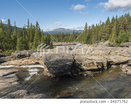 Beautiful northern landscape with rapids and cascades of wild river Kamajokk, boulders and spruce tree forest and hills in Kvikkjokk village in Swedish Lapland. Summer sunny day, blue sky white clouds 84875762