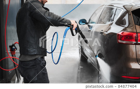 Cropped view of young man washing car on carwash station outdoor. Handsome driver cleaning automobile, using high pressure water. Cropped view of young man washing car on carwash station outdoor. Handsome driver cleaning automobile, using high pressure water. 84877404