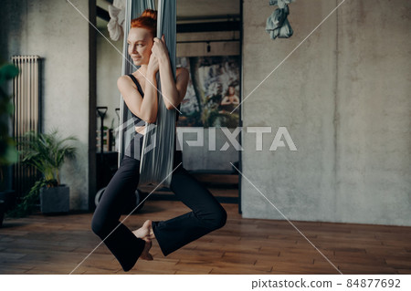 Slender young woman with red hair practices anti-gravity fly yoga Slender young woman with red hair practices anti-gravity fly yoga 84877692