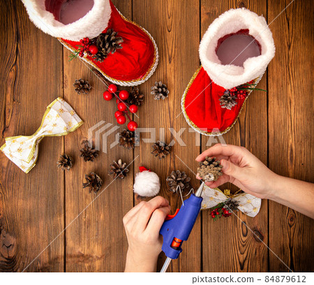 The girl glues a bump on the red boot of santa claus with a glue gun. Making Christmas decorations. Handmade, wooden background 84879612