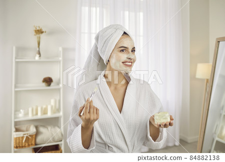 Beauty treatments at home. Beautiful woman in morning applies moisturizing mask or cream on face with brush. Portrait of beautiful woman in bathrobe and towel. Concept of skin care and lifestyle. 84882138