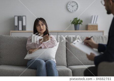 Depressed arab lady sitting on couch, hugging pillow and having consultation with psychologist at clinic, copy space 84882781