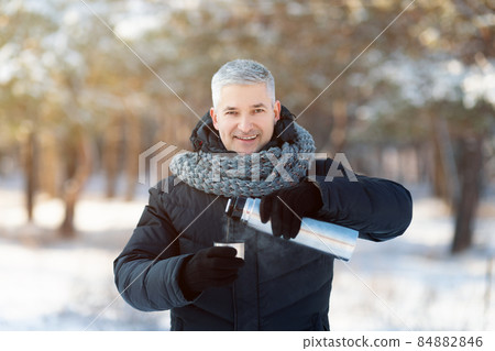 Smiling senior man in warm winter clothes pouring tea from thermos flask in snowy park 84882846