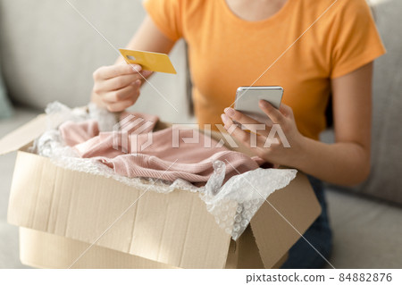 Modern home shopping. Young unrecognizable woman with credit card and phone, unpacking cardboard box with clothes Modern home shopping. Young unrecognizable woman with credit card and phone, unpacking cardboard box with clothes 84882876