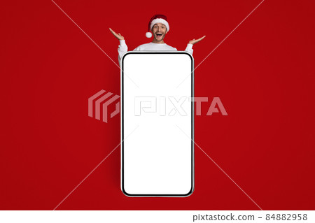 Online Offer. Funny Man In Santa Hat Standing Behind Big Blank Smartphone 84882958