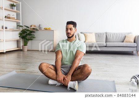 Peaceful millennial Arab guy sitting in lotus pose, meditating with closed eyes on mat at home, copy space 84883144