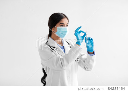 Serious young indian lady doctor in coat, protective mask and gloves with syringe and vaccine on white background 84883464