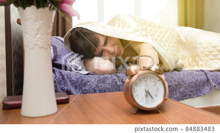 A sleepy, sleepless woman reluctantly reaches out from under the blanket to the alarm clock 84883483