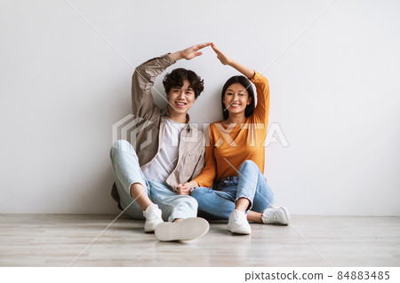 Asian couple making house roof, joining hands above heads as symbol of new home, sitting on floor against white wall 84883485