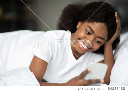 Happy african american woman lying in bed reading text message Happy african american woman lying in bed reading text message 84883488