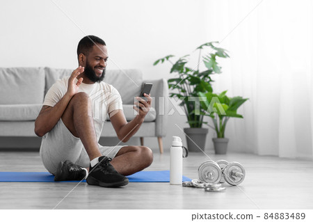 Cheerful young black guy sits on mat on floor in room interior with dumbbells and bottle of water, waves hand Cheerful young black guy sits on mat on floor in room interior with dumbbells and bottle of water, waves hand 84883489