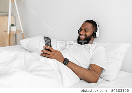 Cheerful young african american man with beard in headphones, typing on smartphone, enjoy listening music 84883506