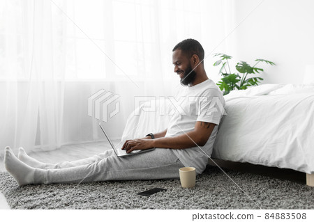 Cheerful young african american man with beard working on laptop in bedroom on floor on window background 84883508