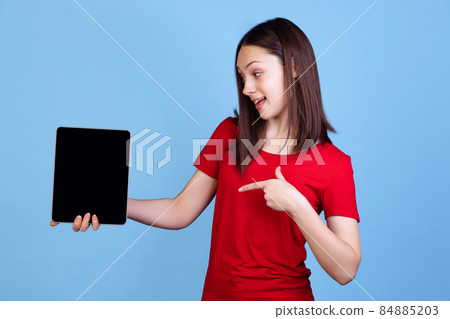Emotional young beautiful girl in red t-shirt using digital tablet isolated on blue studio background. Concept of emotions 84885203