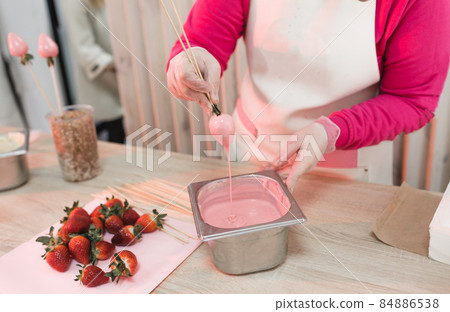 A pastry chef dips strawberries on a wooden skewer into a container with pink chocolate. Preparation of desserts. Chocolate drips from strawberries. A pastry chef dips strawberries on a wooden skewer into a container with pink chocolate. Preparation of desserts. Chocolate drips from strawberries. 84886538
