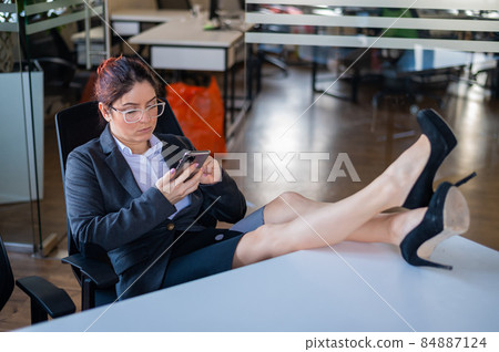 Business woman put feet on work desk and uses mobile phone in office Business woman put feet on work desk and uses mobile phone in office 84887124
