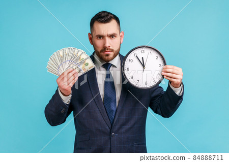 Time to make money. Sad man with beard wearing official style suit holding big clock and dollars banknotes, looking at camera serious facial expression. Indoor studio shot isolated on blue background. 84888711