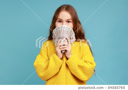 Portrait of little girl hiding half face with money, peeking out of dollar bills, looking at camera, wearing yellow casual style sweater. Indoor studio shot isolated on blue background. 84888768