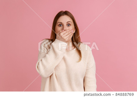 Portrait of shocked blond woman put hand on mouth and looking with fear in her eyes, keeping terrible secret, wearing white sweater. Indoor studio shot isolated on pink background. Portrait of shocked blond woman put hand on mouth and looking with fear in her eyes, keeping terrible secret, wearing white sweater. Indoor studio shot isolated on pink background. 84888769